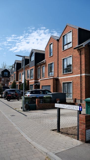 A row of modern, multi-story residential buildings constructed with red brick and black cladding, featuring large windows and pitched roofs, situated along Collier Row Road. In the foreground, there i